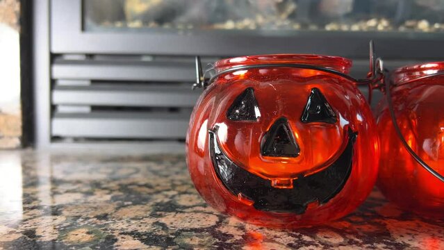 Two bright orange candlesticks in the form of pumpkins Jack-o-lantern Glass lantern with real candles Black eyes and black smile Transparent orange glass Standing in front of a fireplace