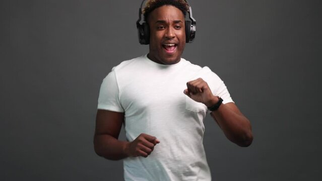 Close-up of an African-American man in a white T-shirt listening to music with headphones and dancing cool. Dancing of a black man on a gray background with headphones