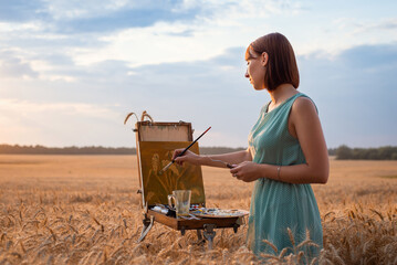Joyful relaxed girl artist painting a picture in the wheat field. Inspired painter works on nature,...