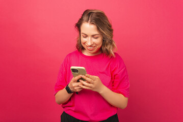 Bright blonde smiling woman wearing pink t-shirt is browsing her phone. Studio shot over magenta background.