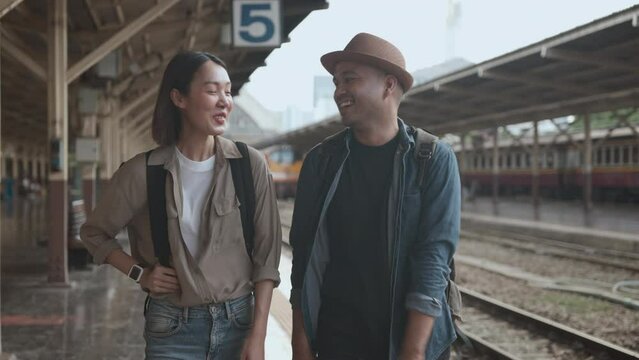 Happy asian handsome man and beautiful woman backpacker wearing hat and backpack walking and talking together in the train station going to travel. 4k resolution.