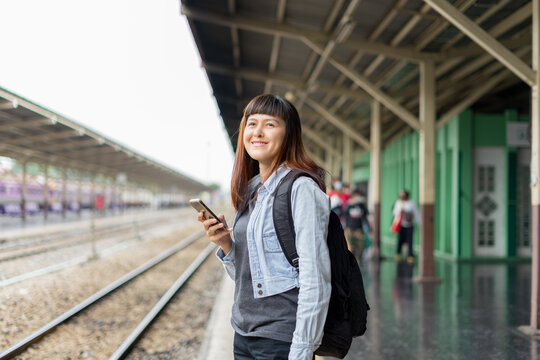 Asian woman waiting the train at train station for travel. Backpack traveler using map on smartphone standing alone at train station platform. Tourist adventure holiday traveling concept.