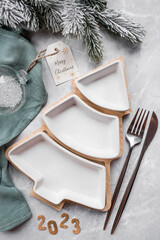 Table setting. A plate in the form of a Christmas tree, a green napkin, spruce branches and Christmas decorations on a gray background. The concept of Merry Christmas, New Year 2023 and restaurants.