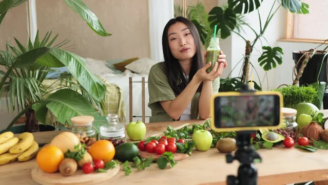 Young asian woman blogger or content creator preparing smoothies and recording video on phone. Attractive female showing how cook healthy food while recording blog with phone at stylish kitchen
