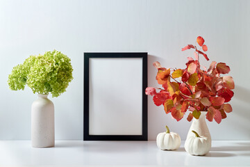 Mockup with a black frame and colorful autumn leaves in a vase on a light background. Empty poster...