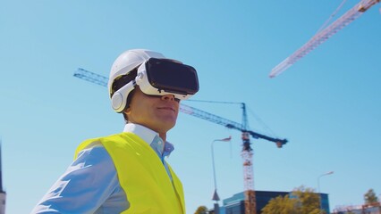 Professional builder in VR helmet standing in front of construction site and using virtual and...