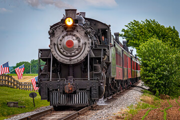 Fototapeta na wymiar View of a Restored Steam Train Approaching Head-On With American Flags Waving on a Fence on a Spring Day
