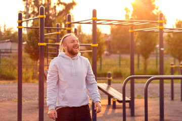 A man with a beard and an interesting hairstyle stands on the sports ground in the morning and smiles.