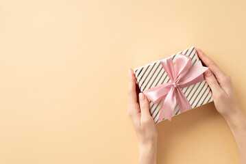 New Year concept. First person top view photo of woman's hands giving present box with pink ribbon bow on isolated pastel beige background with blank space