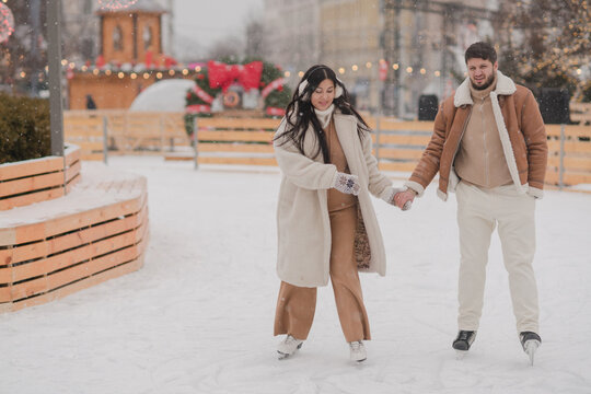 Cheerful loving couple in warm cozy clothes walk outdoors on festive city streets. Man and woman skating on ice rink as snow falls. Cold happy winter day. Holidays, Christmas, New Year, love concept. - Powered by Adobe