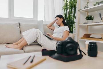 Asian woman photographer lying at home on the couch and reviewing photos from the camera in laptop for color correction retouching, work as a freelance business photographer for yourself home office