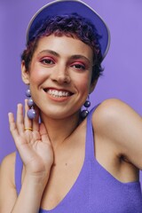Portrait of a sporty fashion woman posing smiling with teeth at the camera with stylish earrings in her ears in a purple yoga tracksuit and a transparent cap on a purple monochrome background