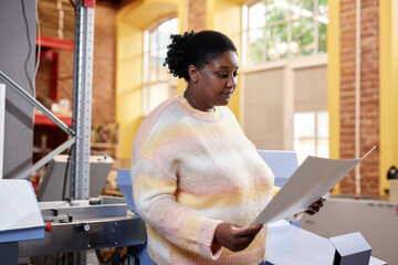 Portrait of young black woman operating printing machine and looking at color test sheet, copy space