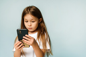 OMG, shocked little schoolgirl holding smartphone on blue background in studio. A little girl...