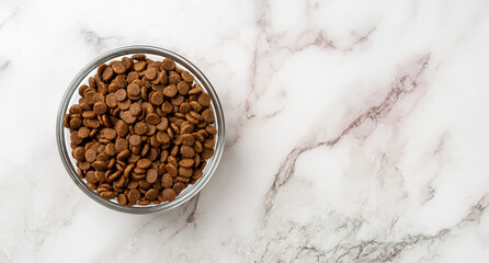 Dry pet food in a glass bowl over marble background. Glass feeding bowl full of gluten free dry protein kibbles for cats closeup. Complete food for domestic animals concept. Copy space.