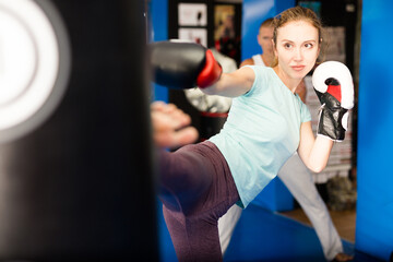 Woman in boxing gloves kicking punchbag during training. Trainer standing behind and watching.