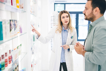 Customer entered in pharmacy store and looking for some medical pills and cremes. Young pharmacist woman is helping him