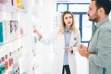 Foto op Canvas Customer entered in pharmacy store and looking for some medical pills and cremes. Young pharmacist woman is helping him © Nemanja