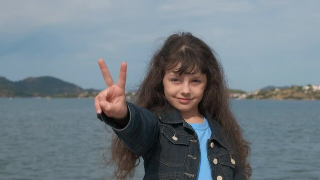 Girl show sign of peace on shore. A playful child show the peace sign against sea water and tropical hills during summer vacation.