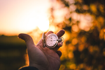 Aufgeklappte Taschenuhr in der Hand gehalten vor einem Bokeh-Hintergrund im Sonnenuntergang