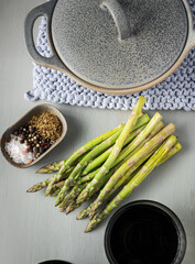 Green asparagus bunch over a wooden table surrounded by a tray of spices and en elegant ceramic pot.