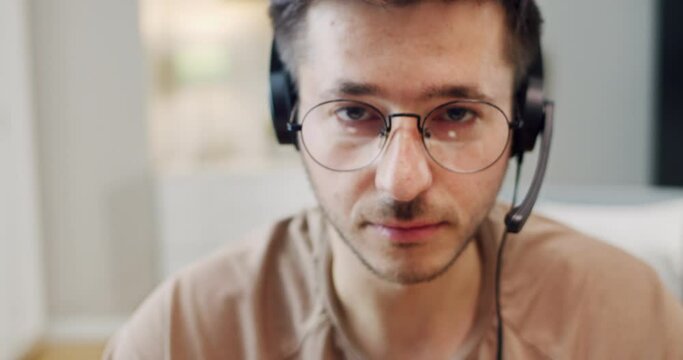 Camera zooming in on face of young Caucasian male worker of client support or call center in headset with mic and glasses. Indoors. Blurred. Man working at home and having videochat.