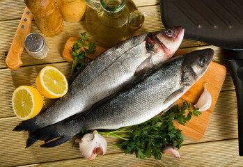 View of branzino fish on cutting board on wooden surface with fresh lemon, parsley, garlic and...