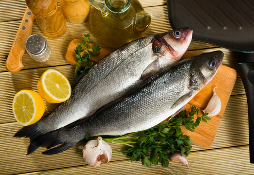 View of branzino fish on cutting board on wooden surface with fresh lemon, parsley, garlic and spices
