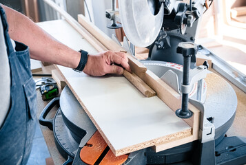 Close-up of carpenter cutting a wooden baseboard. Male carpenter sawing a baseboard with a circular saw in a carpentry workshop close-up. Using a miter saw to cut a piece of wood
