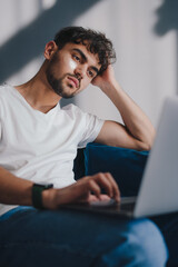 Handsome young Caucasian man, sitting on sofa at home with hydrogel patches under eyes, working on laptop. Digital technology concept. Education internet