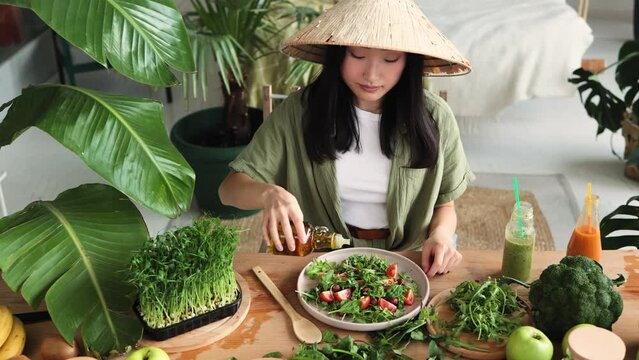 Young attractive asian woman in traditional conical hat sitting at table in stylish light tropical kitchen and pouring olive oil to healthy vegetable salad.