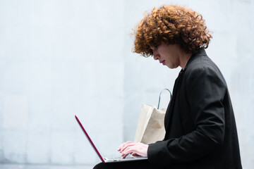 A non-binary person with curly hair in a black suit types on their laptop to make a purchase.