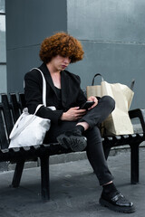 a non-binary person sitting on a bench with shopping bags watching the smartphone