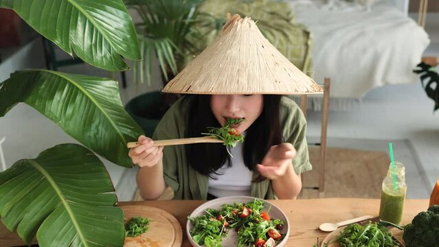 Beautiful young and happy asian woman eating healthy salad with fresh organic vegetables, sitting on table with green fresh ingredients at stylish light exotic studio.
