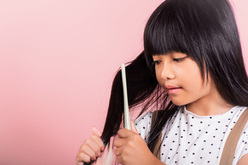 Asian little kid 10 years old hold comb brushing her unruly she touching her long black hair at studio shot isolated on pink background, Happy child girl with a hairbrush, Hair care concept - obrazy, fototapety, plakaty