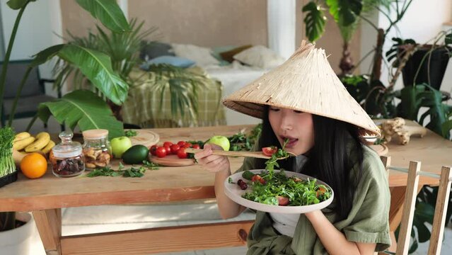 Beautiful happy young asian woman in traditional conical hat holding and eating healthy salad sitting at chair near table with organic vegetables and fruits ingredients indoors at tropical resort.