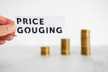 hand holding Price Gouging text in front of growing stacks of coins, symbol of excessive pricing or...