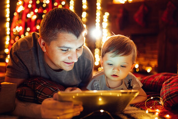 Dad and little son using tablet pc at home near Christmas tree in cozy living room on Christmas eve