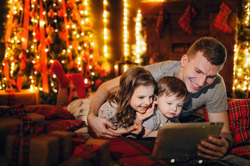 Dad with two cute kids using tablet pc at home by fireplace in cozy living room on Christmas eve