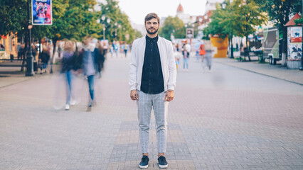 Portrait of young businessman in trendy garments standing in busy pedestrian street and looking at...