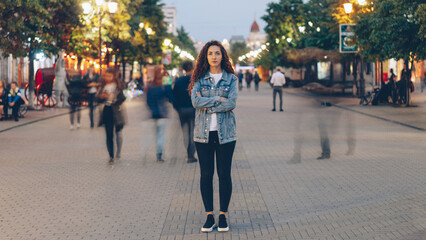 Portrait of stylish young lady tired of usual haste standing in the street among whizzing people and looking at camera. Time, youth and modern society concept. - 542859098