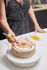 Cropped close-up shot of a woman's hand decorating a cake using a double-sided modeling tool.  A woman decorates the cake on a white turntable on the table in the kitchen. Top view.