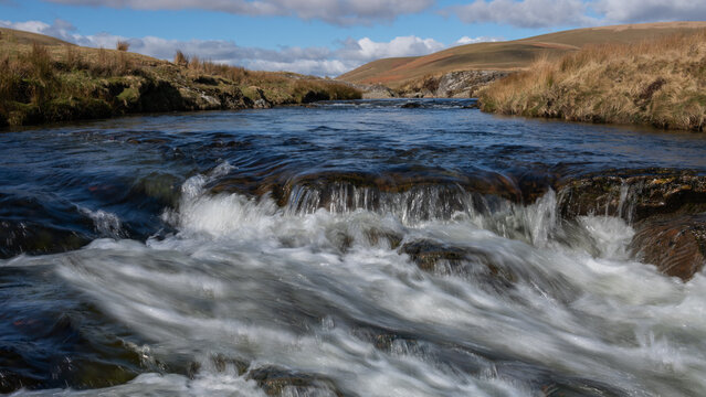 A low level image looking up a small river towards distant hills. It shows small white water rapids in the foreground