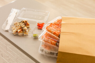 Sushi rolls in plastic box near paper package on white table. Delivery. Take away.