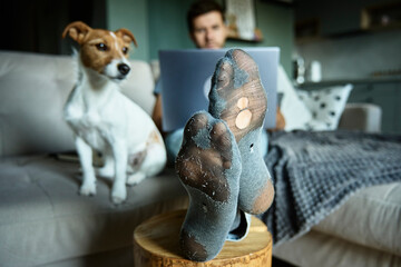 Freelancer work from home. Man in leaky socks using laptop and resting on sofa with his dog