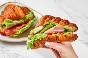 Female hand holding croissant with fried egg, ham, salami, cheese, tomatoes and lettuce