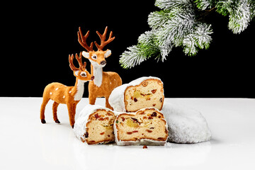 Slices of sweet stollen with nuts and dried fruit on table with toy deers and snow-covered spruce...