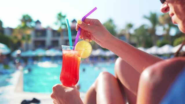Woman sitting on beach chair and holding in hand drinking and stir cocktail with tubule, near at blue swimming pool at background beautifully tanned legs. Relax, rest, vacation. Resort spa hotel. 