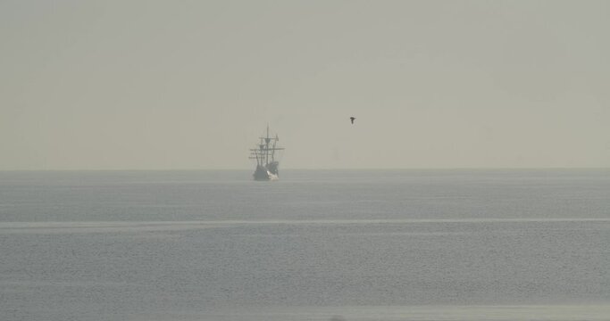 Ferdinand Magellan Nao Victoria carrack boat replica sails in the distance in the mediterranean at sunrise while a bird flies in calm sea in slow motion 60fps