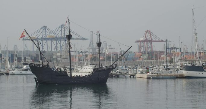 Ferdinand Magellan Nao Victoria carrack boat replica with spanish flag enters wharf in Valencia with containers and cranes in the background in slow motion 60fps